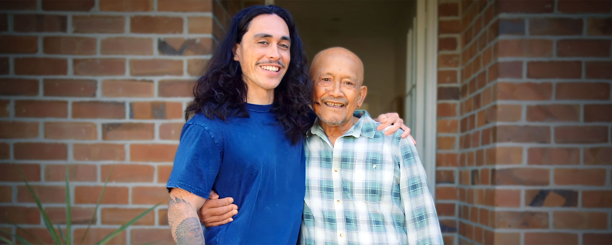 A young 20 something man proudly stands with a protective arm around his granddad.