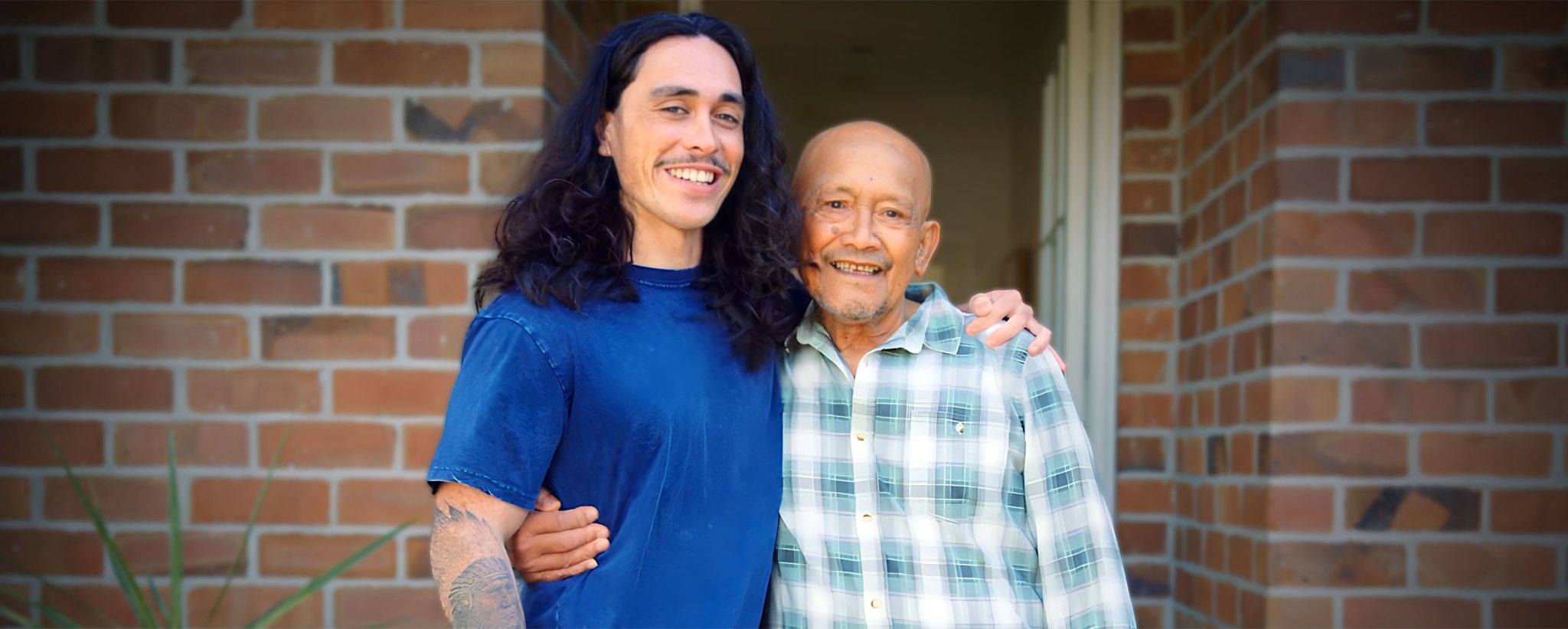 A young 20 something man proudly stands with a protective arm around his granddad.
