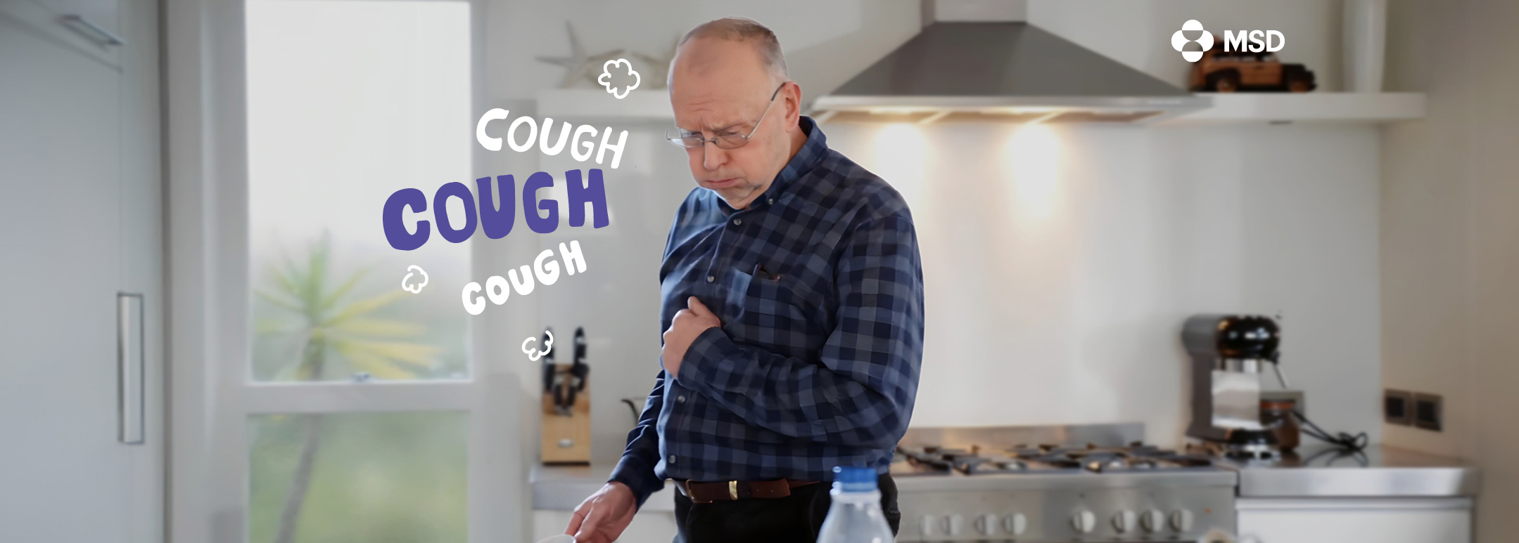 A mid to older aged man is coughing in his kitchen.